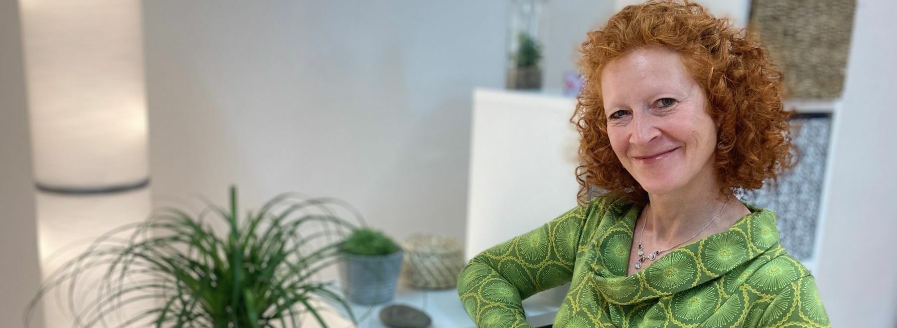 Lächelnde Frau mit lockigem rotem Haar in einem grünen Oberteil, drinnen in der Nähe von Zimmerpflanzen.