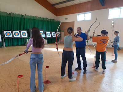Fünf Personen üben Bogenschießen im Innenbereich und zielen mit ihren Bögen auf Ziele an der Wand.