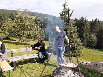 Zwei Männer kochen Essen über einem Lagerfeuer in einem grasbewachsenen Waldgebiet.