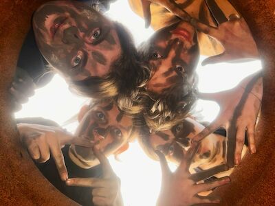 Vier Personen mit bemalten Gesichtern blicken in eine runde Öffnung hinunter und formen Handbewegungen.