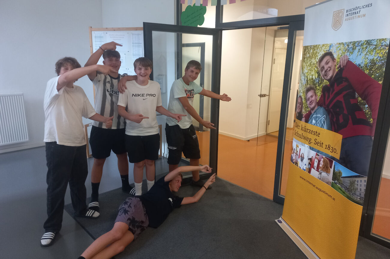 4 Schüler stehen vor der geöffneten Eingangstür zum Internat, einer liegt am Boden und alle machen gemeinsam eine einladende Geste um zum Eintreten ins Internat einzuladen. Neben der Eingangstür steht ein Rollup des Internats.